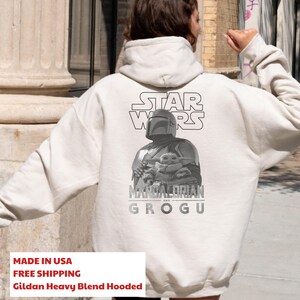 Może przedstawiać: Kremowa bluza z kapturem z czarno-białą grafiką na plecach. Wz&oacute;r przedstawia logo Star Wars nad ilustracją Mandalorianina trzymającego Grogu, z napisem Mandalorian and Grogu wydrukowanym poniżej.