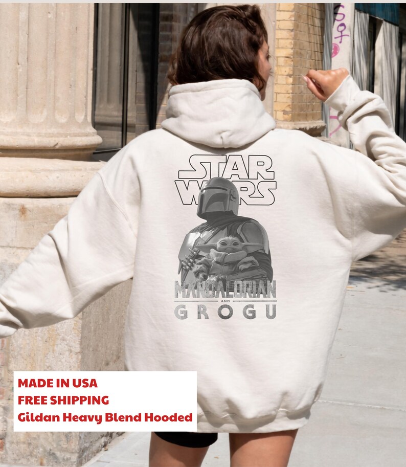 Może przedstawiać: Kremowa bluza z kapturem z czarno-białą grafiką na plecach. Wz&oacute;r przedstawia logo Star Wars nad ilustracją Mandalorianina trzymającego Grogu, z napisem Mandalorian and Grogu wydrukowanym poniżej.