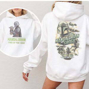 May include: A white hooded sweatshirt featuring a graphic of The Mandalorian and Grogu on the back with the text The Mandalorian and Grogu and This Is The Way. A circular inset shows the front of the hoodie with a smaller graphic and text.