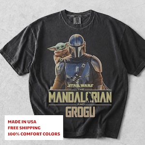 May include: A black short-sleeve t-shirt featuring a graphic print of The Mandalorian and Grogu. The design includes the text Star Wars The Mandalorian and Grogu in a tan font. The shirt has a crew neckline and a faded, vintage-style fabric finish.