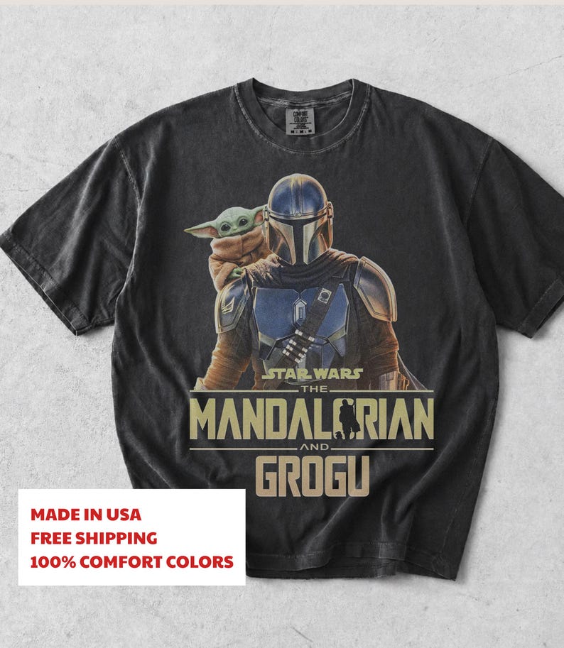 May include: A black short-sleeve t-shirt featuring a graphic print of The Mandalorian and Grogu. The design includes the text Star Wars The Mandalorian and Grogu in a tan font. The shirt has a crew neckline and a faded, vintage-style fabric finish.