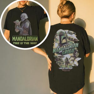 May include: A black t-shirt featuring a graphic print of The Mandalorian and Grogu. The back displays a large collage of the characters with the text "The Mandalorian and Grogu" and "This is the way." A circular inset shows the front design.