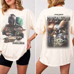 May include: An off-white t-shirt featuring a graphic print of The Mandalorian and Grogu. The front shows the characters with a spaceship and planet, while the back displays a larger action scene with the text Star Wars The Mandalorian.