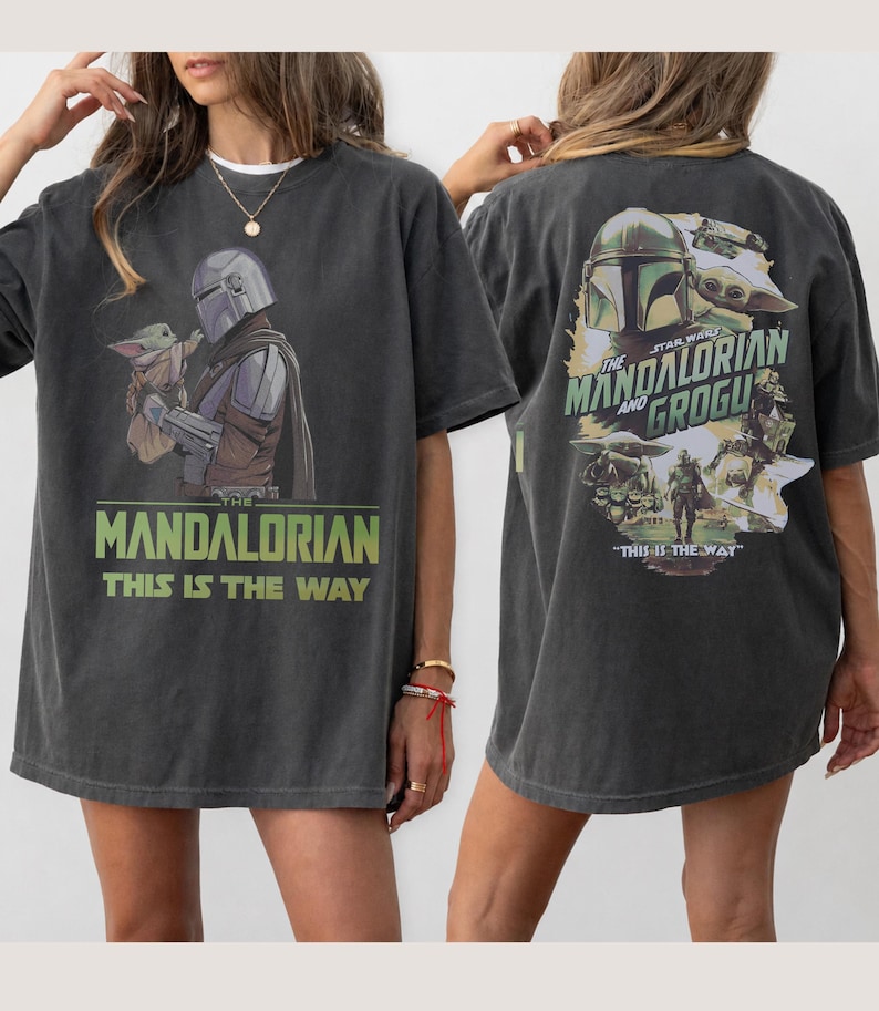 May include: A charcoal gray short-sleeve t-shirt featuring The Mandalorian and Grogu graphics. The front displays the characters with the text "THE MANDALORIAN THIS IS THE WAY". The back shows a collage of scenes with "THE MANDALORIAN AND GROGU".