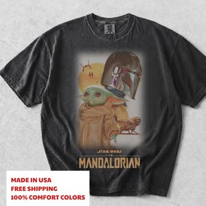 May include: A black short-sleeve t-shirt featuring a graphic print of the Mandalorian helmet and Grogu. The design includes a yellow sun, small spacecraft, and the text Star Wars The Mandalorian in a tan font centered below the characters.