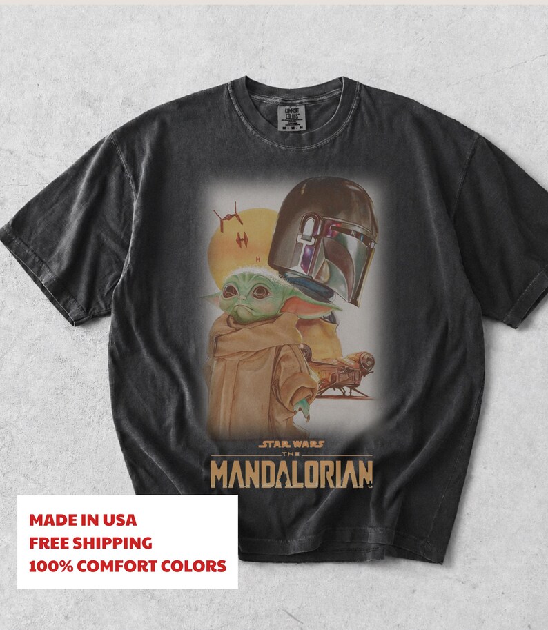 May include: A black short-sleeve t-shirt featuring a graphic print of the Mandalorian helmet and Grogu. The design includes a yellow sun, small spacecraft, and the text Star Wars The Mandalorian in a tan font centered below the characters.