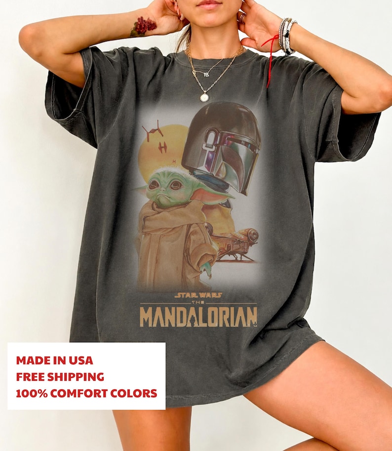 May include: A charcoal gray short-sleeve t-shirt featuring a graphic print of a green creature in a tan robe, a metallic helmet, and a yellow sun. The text STAR WARS THE MANDALORIAN is printed in gold below the graphic on the front of the shirt.