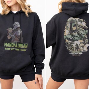 May include: A black hooded sweatshirt featuring a graphic of The Mandalorian and Grogu on the front with the text "Mandalorian This Is The Way". The back displays a larger, detailed illustration of the characters with "The Mandalorian and Grogu" text.