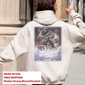 May include: A cream-colored hooded sweatshirt featuring a large graphic on the back. The print depicts characters from The Mandalorian and Grogu, including the Mandalorian, Grogu, and Jabba the Hutt, with the text "The Mandalorian and Grogu Now Playing".