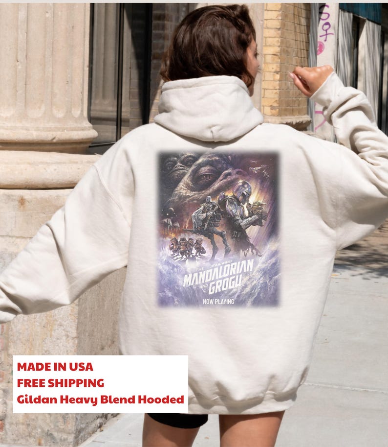 May include: A cream-colored hooded sweatshirt featuring a large graphic on the back. The print depicts characters from The Mandalorian and Grogu, including the Mandalorian, Grogu, and Jabba the Hutt, with the text "The Mandalorian and Grogu Now Playing".