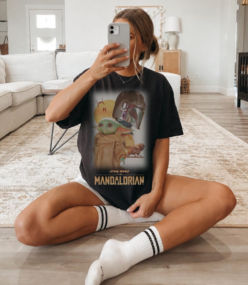 May include: A black short-sleeve t-shirt featuring a graphic print of The Mandalorian and Grogu. The design includes a yellow sun, a helmet, and the text Star Wars The Mandalorian in gold lettering centered on the front of the garment.