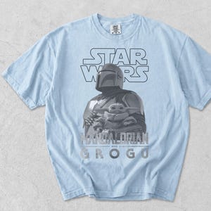 Może przedstawiać: Jasnoniebieski t-shirt z kr&oacute;tkim rękawem z nadrukiem graficznym przedstawiającym Mandalorianina i Grogu. Wz&oacute;r zawiera napis STAR WARS u g&oacute;ry oraz MANDALORIAN AND GROGU u dołu szarą czcionką. Koszulka posiada okrągły dekolt.
