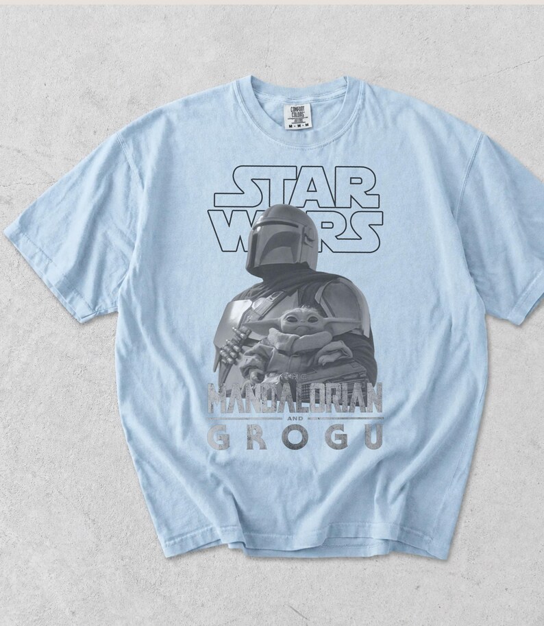 Może przedstawiać: Jasnoniebieski t-shirt z kr&oacute;tkim rękawem z nadrukiem graficznym przedstawiającym Mandalorianina i Grogu. Wz&oacute;r zawiera napis STAR WARS u g&oacute;ry oraz MANDALORIAN AND GROGU u dołu szarą czcionką. Koszulka posiada okrągły dekolt.