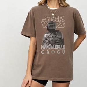 Może przedstawiać: Brązowy t-shirt z kr&oacute;tkim rękawem z nadrukiem graficznym przedstawiającym Mandalorianina i Grogu. Projekt zawiera napis STAR WARS u g&oacute;ry oraz THE MANDALORIAN AND GROGU u dołu białymi literami, wraz z monochromatycznym obrazem postaci.