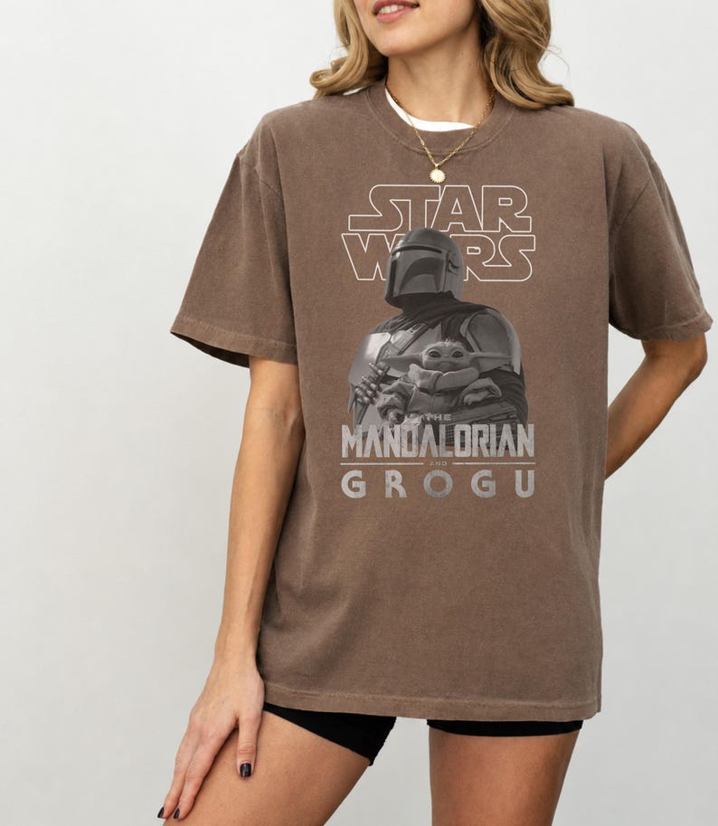 Może przedstawiać: Brązowy t-shirt z kr&oacute;tkim rękawem z nadrukiem graficznym przedstawiającym Mandalorianina i Grogu. Projekt zawiera napis STAR WARS u g&oacute;ry oraz THE MANDALORIAN AND GROGU u dołu białymi literami, wraz z monochromatycznym obrazem postaci.