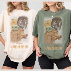 May include: Two short-sleeve t-shirts, one cream and one sage green, each featuring a graphic print of The Mandalorian and Grogu. The design includes a yellow sun, a spacecraft, and the text Star Wars The Mandalorian in gold lettering.