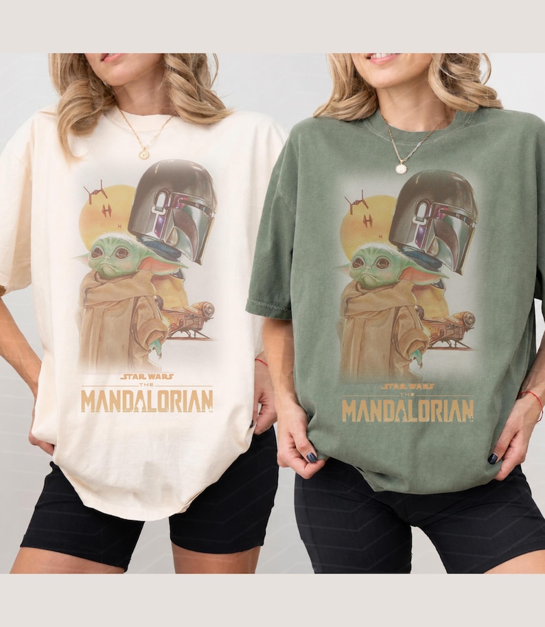 May include: Two short-sleeve t-shirts, one cream and one sage green, each featuring a graphic print of The Mandalorian and Grogu. The design includes a yellow sun, a spacecraft, and the text Star Wars The Mandalorian in gold lettering.