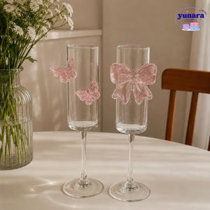 May include: Two clear champagne flutes with pink decorations. One flute has two pink butterfly adornments, the other a pink bow. The flutes are on a white surface, with a vase of flowers in the background.