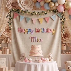 May include: A birthday party setup featuring a beige fabric backdrop with the text Happy birthday Mia, a two-tier cake, cupcakes, and teddy bears. The scene is decorated with a balloon arch in gold, pink, and blue, paper flowers, and string lights.