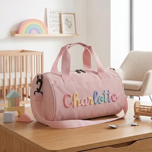 May include: A cylindrical pink corduroy duffel bag featuring the name Charlotte embroidered in multi-colored yarn letters on the front. The bag includes two top handles, a detachable shoulder strap, and a zippered closure with black hardware.