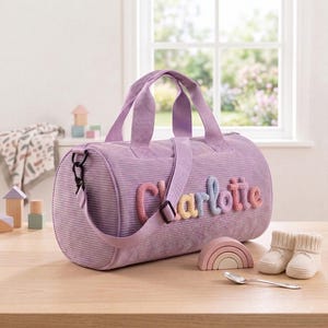 May include: A lavender corduroy duffel bag featuring the name Charlotte in raised, multi-colored knit lettering on the side. The bag includes two top handles, a detachable shoulder strap with a black buckle, and a zipper closure along the top.
