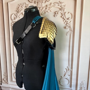 May include: A shoulder armour piece featuring gold-coloured metallic scales attached to a black leather strap system with a silver-toned metal ring. A long, flowing teal-coloured fabric cape is draped from the shoulder harness assembly.