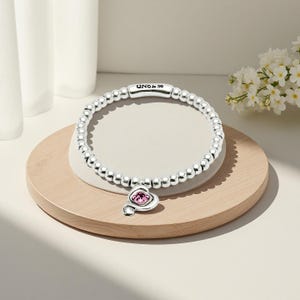 Peut inclure: Un bracelet en argent compos&eacute; de perles avec un charm en forme de c&oelig;ur orn&eacute; d'une pierre rose. Le bracelet porte le nom de la marque "UNO & 50". Il est pr&eacute;sent&eacute; sur une surface en bois clair.