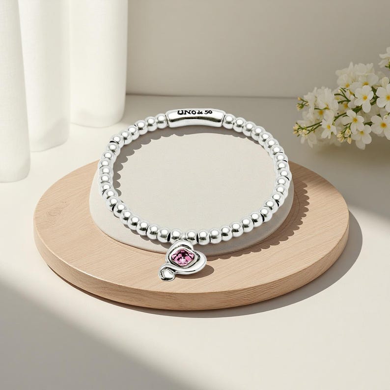 Peut inclure: Un bracelet en argent compos&eacute; de perles avec un charm en forme de c&oelig;ur orn&eacute; d'une pierre rose. Le bracelet porte le nom de la marque "UNO & 50". Il est pr&eacute;sent&eacute; sur une surface en bois clair.