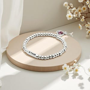 Peut inclure: Un bracelet de perles argent&eacute;es avec un charm en pierre rose. Le bracelet comporte une barre cylindrique avec le texte "UNOde50". Il est pr&eacute;sent&eacute; sur une plateforme circulaire beige, avec des fleurs blanches en arri&egrave;re-plan.