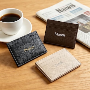 Könnte beinhalten: Drei personalisierte Leder-Kartenetuis in Schwarz, Braun und Beige, jeweils mit einem eingeprägten Namen. Das schwarze Kartenetui hat "Phelan" in Gold, das braune "Maren" in Weiß und das beige "Jorah" in Silber. Eine Tasse Kaffee und eine Zeitung sind im Hintergrund.