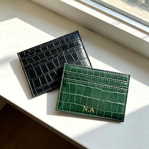 May include: Two rectangular card holders with a crocodile-embossed texture. One card holder is black, and the other is dark green with the gold-coloured initials N.A. printed on the bottom centre. Both items feature multiple slots for cards.