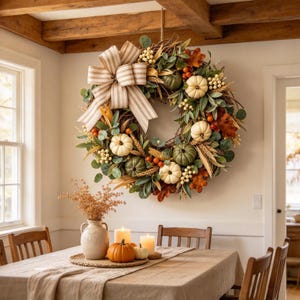May include: A round wreath featuring a large beige and white striped bow, small white and green pumpkins, orange berries, wheat stalks, and green leaves on a twig base. The wreath is decorated with autumn-themed foliage and natural textures.