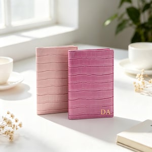 May include: Two rectangular passport holders with a textured crocodile skin pattern. One holder is light pink, and the other is a darker magenta pink with the gold embossed letters DA in the bottom right corner. Both are standing upright.