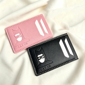 May include: A pink and a black rectangular card holder, each featuring a debossed graphic of a masked figure and the text CAT ID. The card holders have white stitching along the edges and two vertical white slots for holding identification cards.