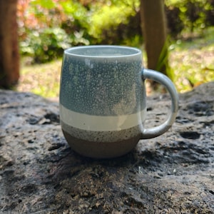 May include: A ceramic mug with a rounded shape and a handle. The exterior features a horizontal color-blocked design with a speckled light blue top section, a solid white middle band, and a speckled gray-brown base with a natural clay finish.