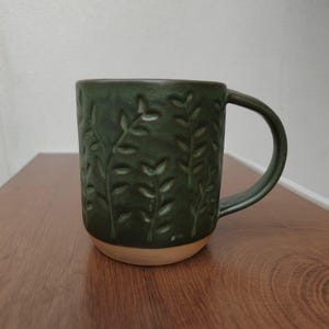 May include: A dark green ceramic mug featuring a debossed leaf pattern across its exterior surface. The mug has a smooth, curved handle and a contrasting unglazed light beige base, showcasing a textured finish with vertical plant motifs.