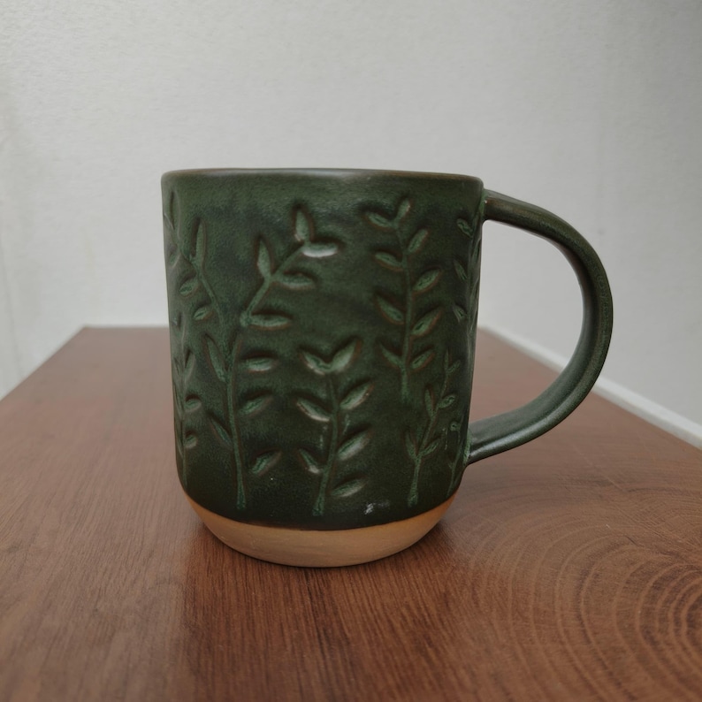 May include: A dark green ceramic mug featuring a debossed leaf pattern across its exterior surface. The mug has a smooth, curved handle and a contrasting unglazed light beige base, showcasing a textured finish with vertical plant motifs.