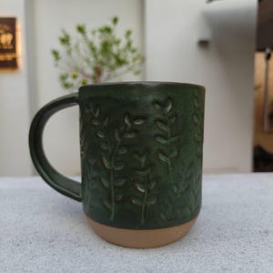 May include: A dark green ceramic mug featuring a textured, embossed pattern of vertical leafy vines. The mug has a curved handle on the left side and a smooth, unglazed light brown base that contrasts with the deep green glazed upper section.