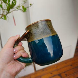 May include: A ceramic mug featuring a two-tone glaze with a deep blue glossy finish on the lower section and a textured, speckled olive-brown finish on the upper rim. The mug includes a curved handle that matches the blue glaze of the base.