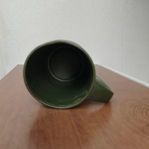 May include: A dark olive green ceramic mug viewed from directly above, showing the circular opening and the interior basin. The mug features a smooth, glossy finish on the exterior and a matching green interior with a sturdy handle attached.