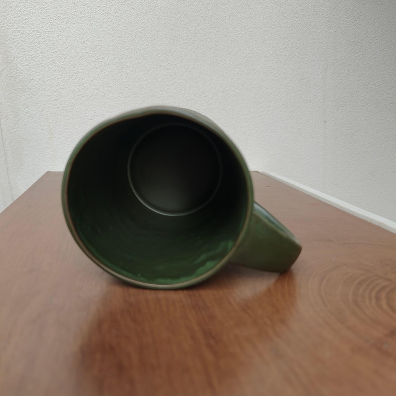 May include: A dark olive green ceramic mug viewed from directly above, showing the circular opening and the interior basin. The mug features a smooth, glossy finish on the exterior and a matching green interior with a sturdy handle attached.