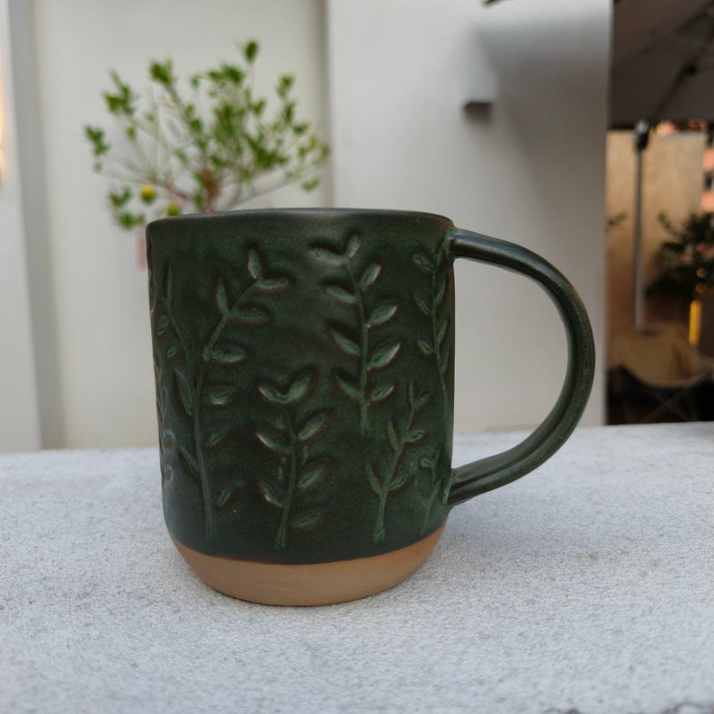 May include: A dark green ceramic mug featuring a textured, embossed pattern of vertical leafy branches. The mug has a smooth, curved handle on the right side and a contrasting unglazed, light tan base that exposes the natural clay material.