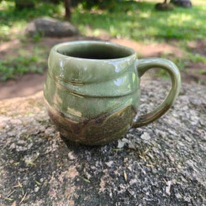 May include: A rounded ceramic mug featuring a glossy sage green glaze on the upper portion that transitions into a textured, earthy brown finish at the base. The mug has a curved handle and a smooth, slightly tapered rim with visible swirling patterns.