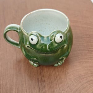 May include: A round ceramic mug shaped like a green frog with a glossy finish. The mug features two protruding eyes on the rim, a wide smiling mouth, and small front feet at the base. The interior is a speckled off-white colour with a handle.