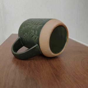May include: A dark green ceramic mug featuring a textured leaf pattern embossed on the exterior surface. The mug has a curved handle and a wide, unglazed light tan base that contrasts with the deep green glaze covering the rest of the body.