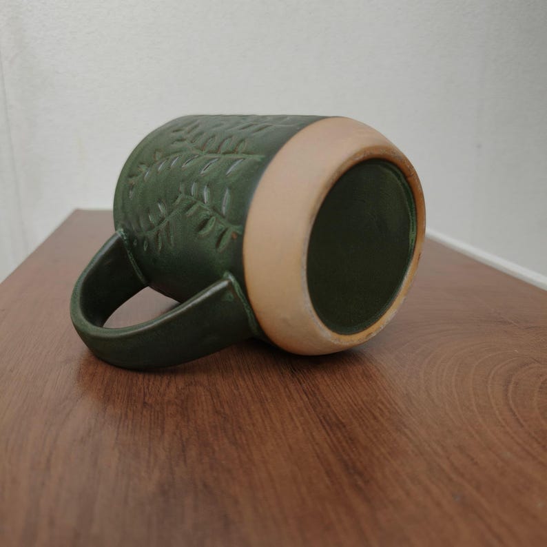 May include: A dark green ceramic mug featuring a textured leaf pattern embossed on the exterior surface. The mug has a curved handle and a wide, unglazed light tan base that contrasts with the deep green glaze covering the rest of the body.
