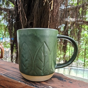 May include: A forest green ceramic mug featuring a repeating embossed leaf pattern around the exterior. The mug has a smooth, dark green glazed finish on the upper portion and a contrasting unglazed, light beige base with a curved handle.