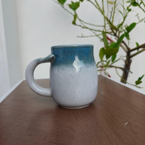 May include: A ceramic mug with a rounded body and a curved handle. The upper portion of the mug features a deep blue glaze, while the lower portion is a light gray with dark speckles throughout the surface, creating a distinct two-tone appearance.