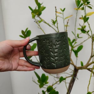 May include: A dark forest green ceramic mug featuring a textured, embossed botanical leaf pattern across the exterior surface. The mug has a smooth, rounded handle and a contrasting unglazed, light tan base at the bottom of the vessel.