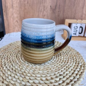 May include: A ceramic mug with a ribbed, textured surface featuring a gradient color scheme. The top section is white, transitioning into a vibrant blue, a deep dark blue, and finally a warm tan base. The handle is glazed in a dark brown tone.